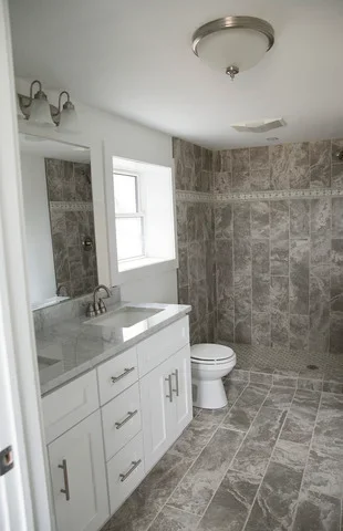 Renovated bathroom with white vanity, granite countertop, and gray tile shower and flooring